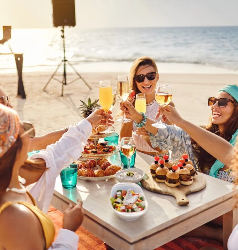 Group of friends enjoying a sandbank meal by the ocean, with gourmet dishes and tropical drinks, with a DJ playing in the background