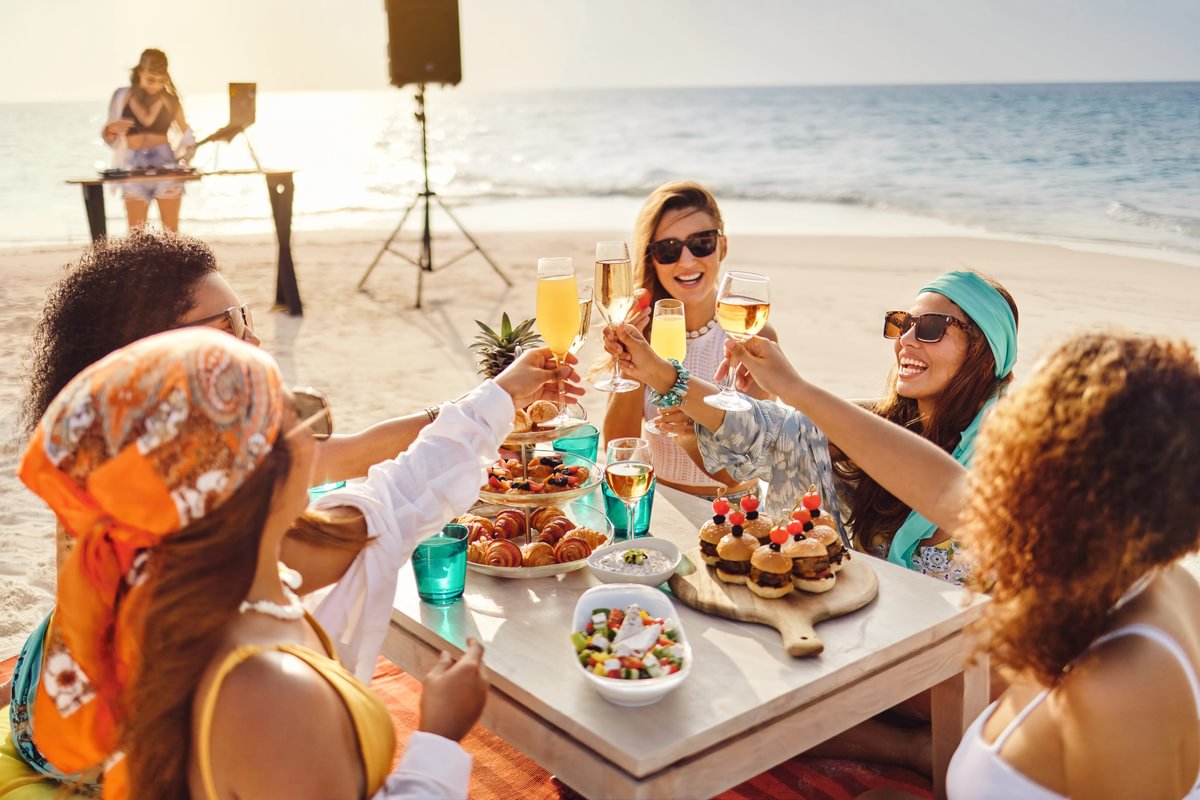 Group of friends enjoying a sandbank meal by the ocean, with gourmet dishes and tropical drinks, with a DJ playing in the background