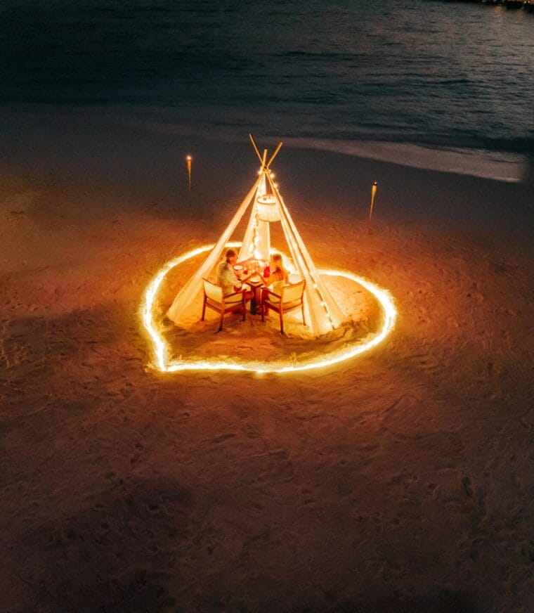 Heart-shaped dining setup on the beach, glowing under the night sky, perfect for a romantic evening.