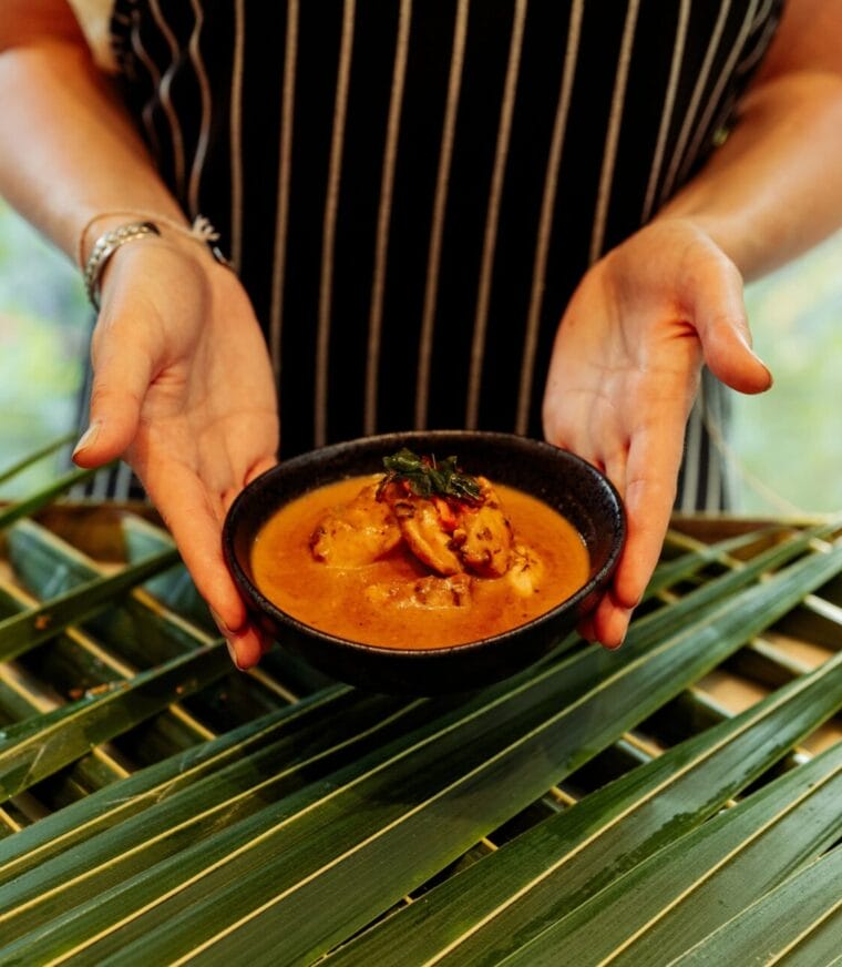 Person holding a black bowl of curry garnished with herbs, with green palm leaves in the background, creating a tropical presentation.