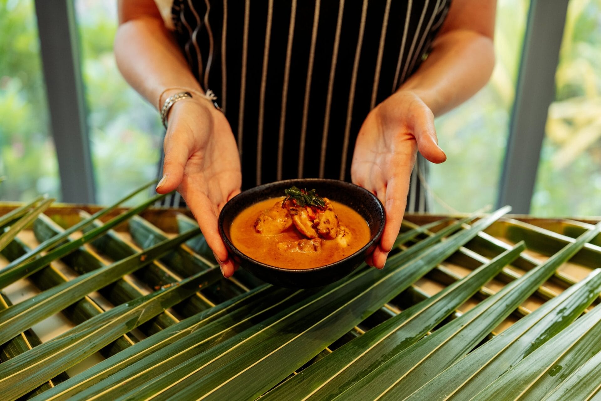 Person holding a black bowl of curry garnished with herbs, with green palm leaves in the background, creating a tropical presentation.