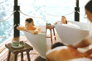 A woman relaxing in a bathtub on an outdoor deck, with the ocean visible in the background. She has a flower in her hair and is enjoying a soothing bath, while a wooden table next to her holds a tea set. The peaceful setting evokes a sense of relaxation and indulgence.