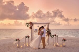 A couple standing under a decorated wedding arch on a sandy beach at sunset. The bride is wearing a white dress and holding a bouquet, while the groom is in a white shirt and tan pants. The arch is adorned with flowers, fabric, and lanterns, creating a dreamy and intimate setting.